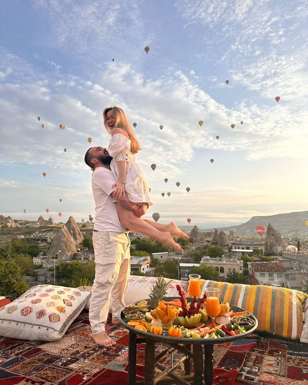 Doors of Cappadocia'nın çatı terasında bir çift, meyve tabağı ve kilim yastıkların yanında; gökyüzü Göreme üzerinde sıcak hava balonlarıyla dolu