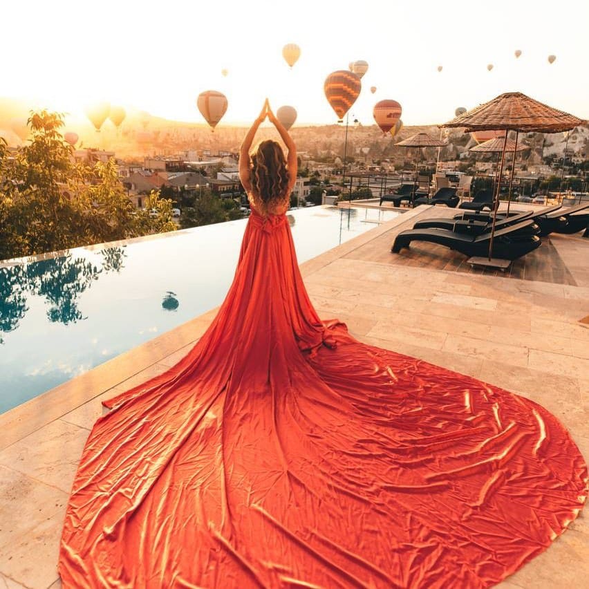 Doors of Cappadocia'nın infinity havuzunun kenarında kırmızı elbiseli bir misafir, gün doğumunda Göreme üzerinde yükselen sıcak hava balonları