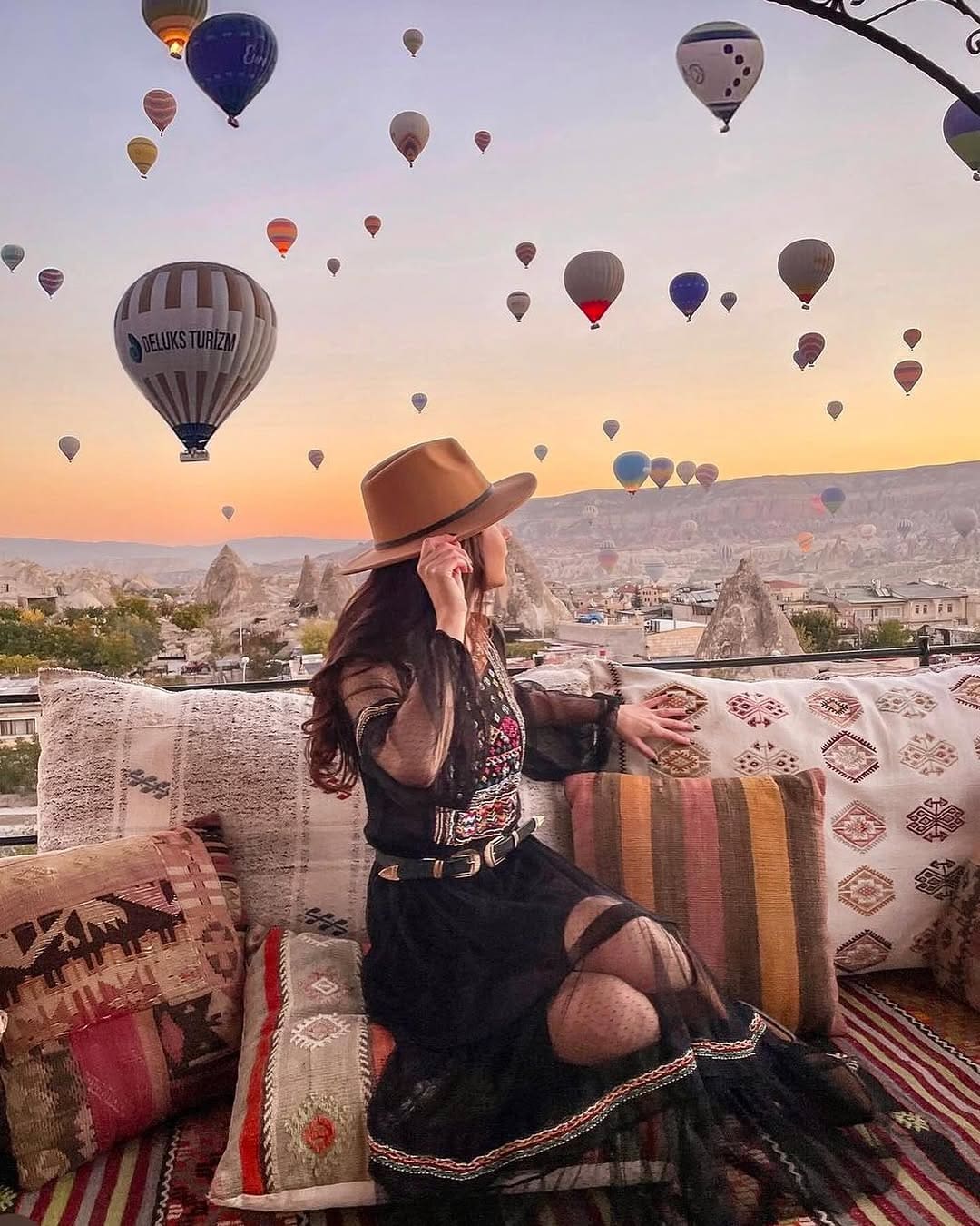 Doors of Cappadocia'da geniş kenarlı şapkalı bir misafir kilim yastıkların üzerinde, gün doğumunda Göreme üzerinde yükselen sıcak hava balonlarını izliyor
