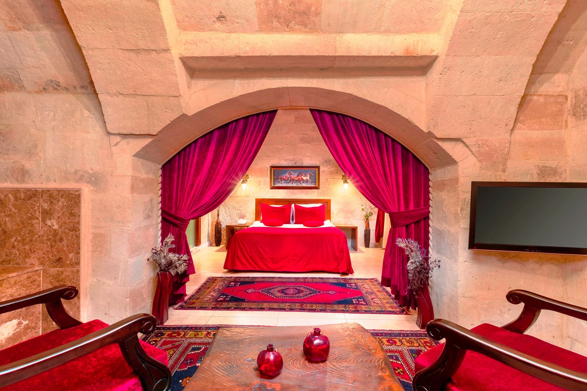 Room 110, Queen Suite: Bed alcove with red curtains under a stone arch, wall-mounted TV.