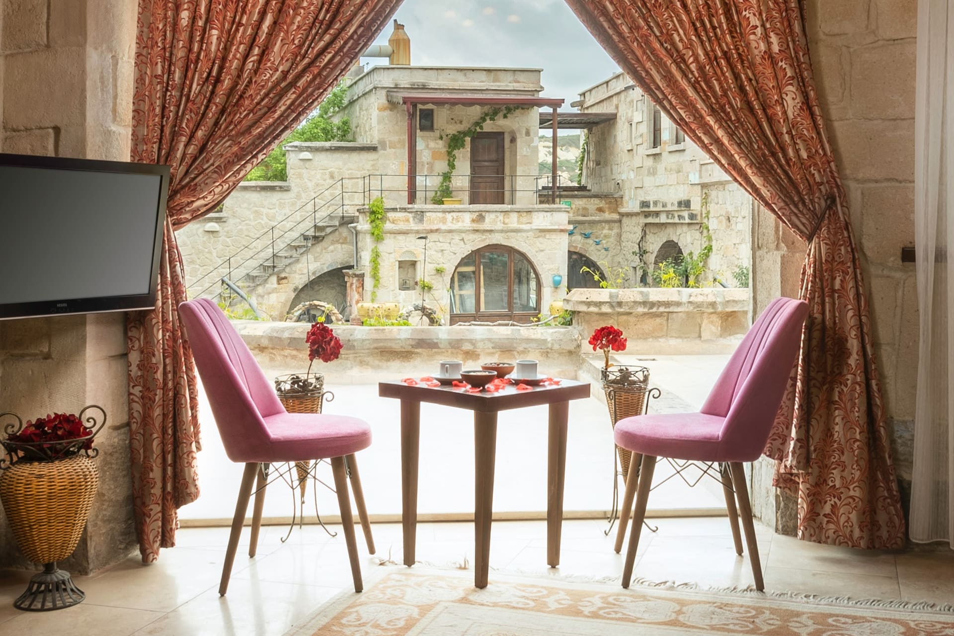Room 111, King Cave Suite: the doorway open to the terrace, pink chairs and a small table, a wall TV, a stone building outside