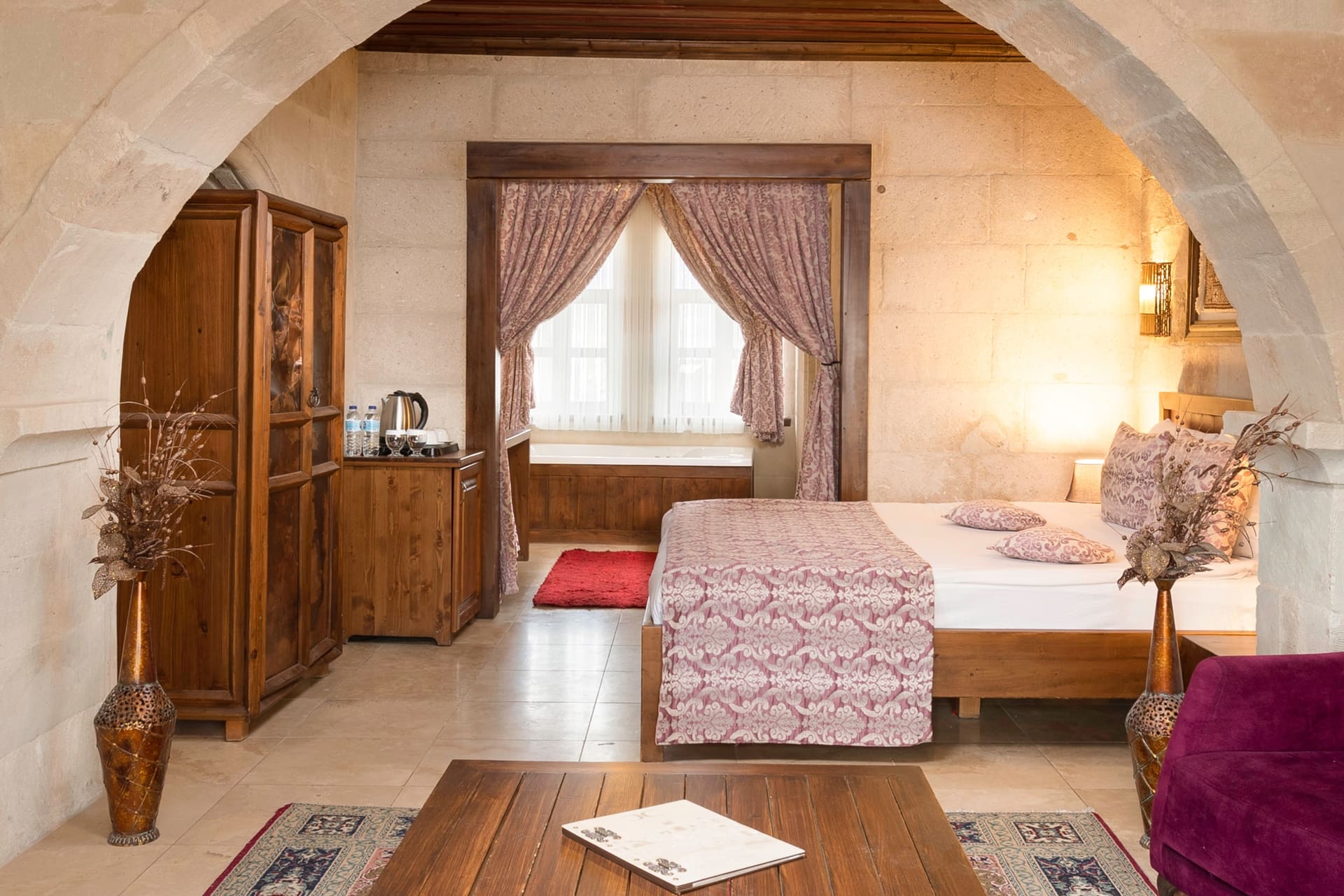 Room 105, King Suite: bed by the curtained window with a purple armchair and a dried flower vase
