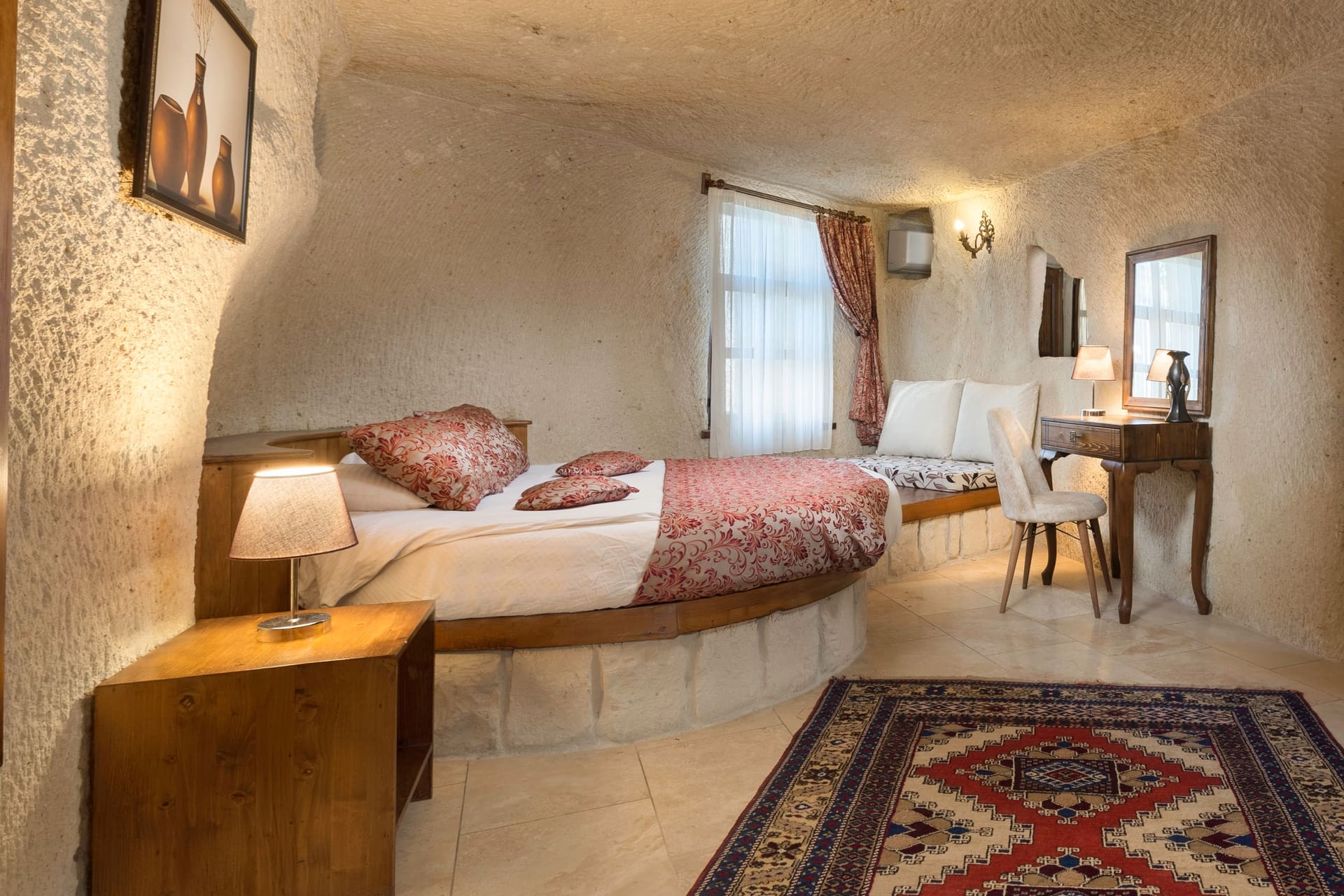 Room 112, King Cave Suite: the pillowed bed, a window with curtains, a bench seat, and a dressing table with mirror
