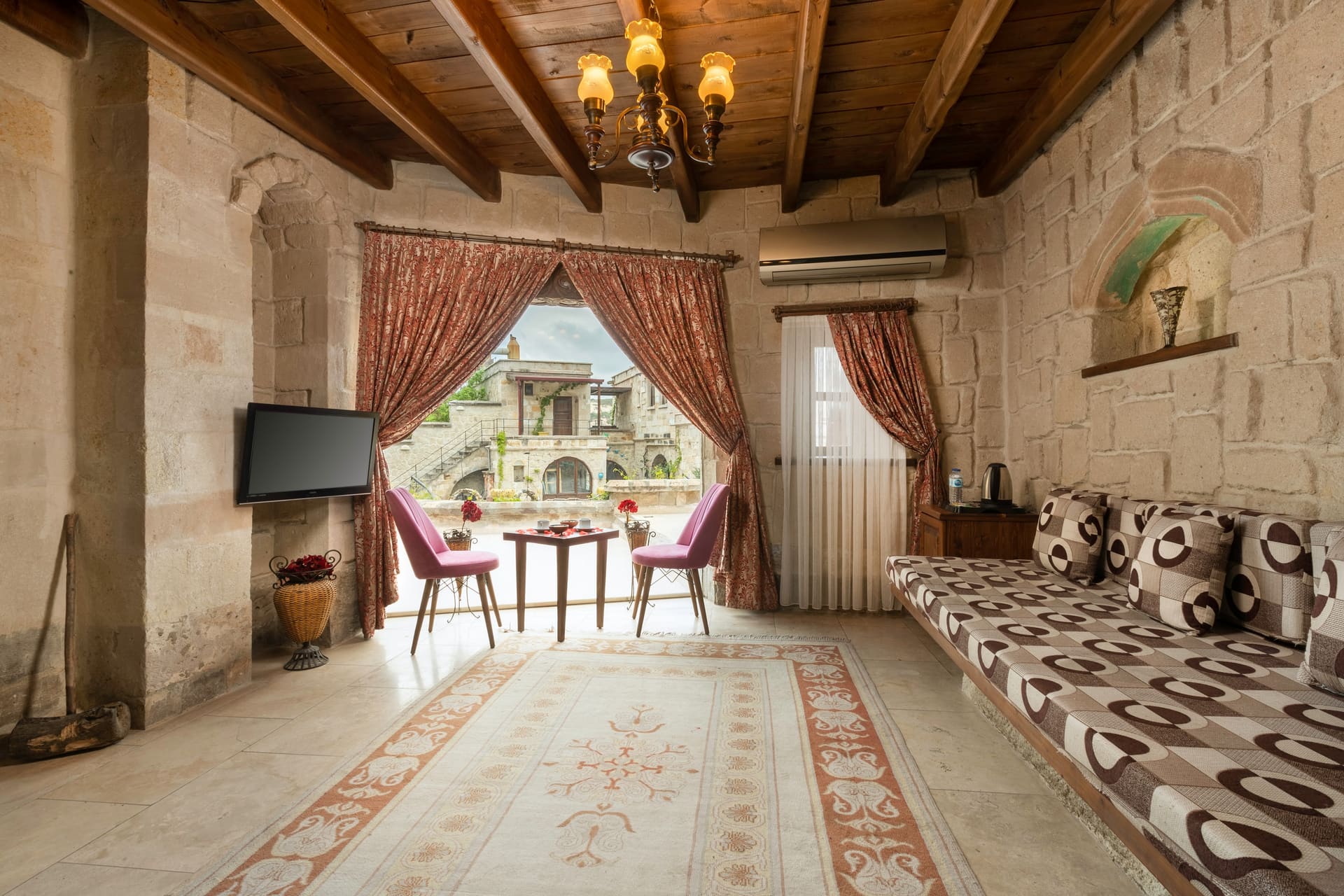 Room 111, King Cave Suite: the wood-beamed ceiling, a chandelier, a patterned bench, and the doorway to the terrace