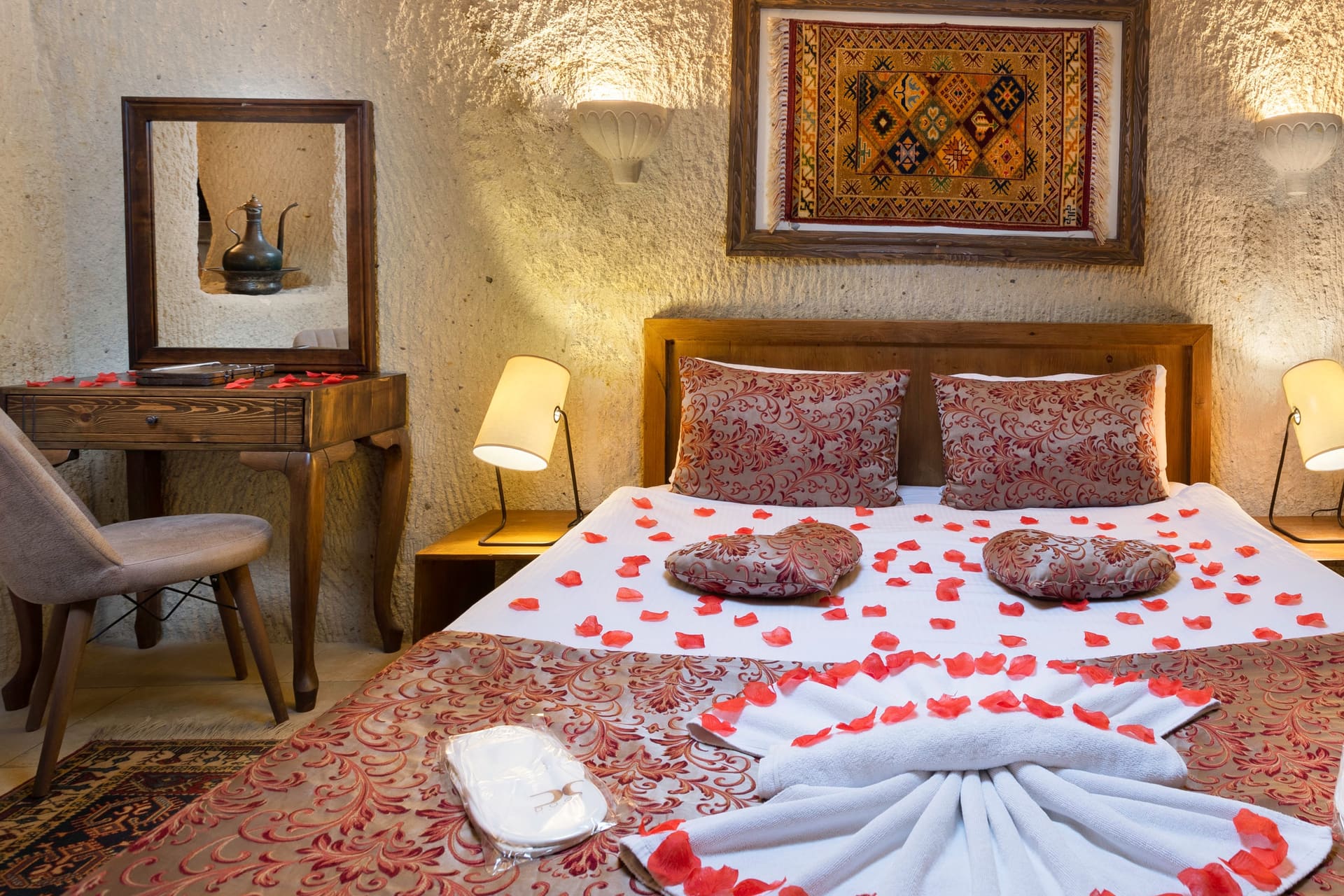 Room 111, King Cave Suite: wide view of the bed with heart-shaped rose petal arrangements, a woven rug artwork on the wall, two bedside lamps